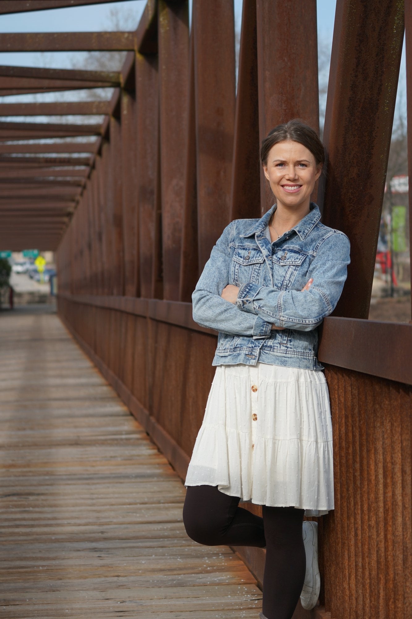 Maria on a rusty brown metal bridge in Batavia, IL, wearing a light blue denim jacket, a white pleated skirt, and black tights.